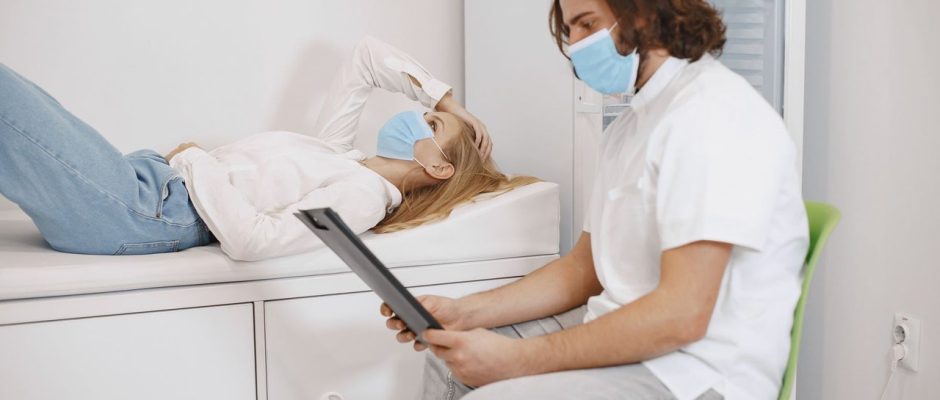 Médico conversa com paciente usando máscara em uma sala de atendimento, enquanto discutem o plano de cuidados.