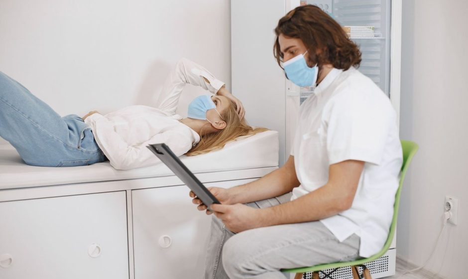 Médico conversa com paciente usando máscara em uma sala de atendimento, enquanto discutem o plano de cuidados.