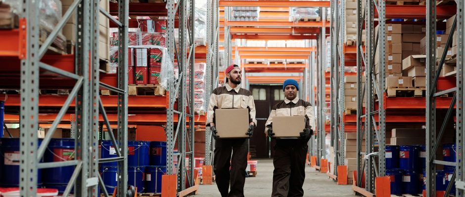 Dois trabalhadores carregam caixas entre prateleiras metálicas em um armazém logístico