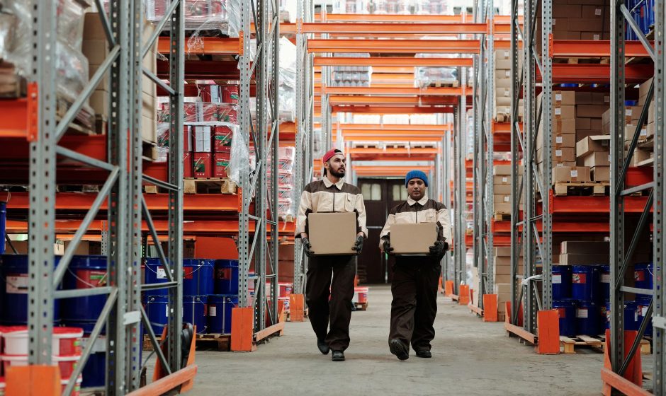Dois trabalhadores carregam caixas entre prateleiras metálicas em um armazém logístico